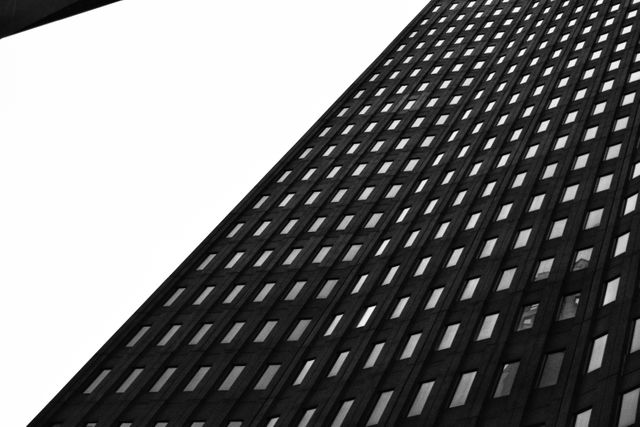 Monochrome image showcasing a tall modern skyscraper with numerous windows against a clear sky, highlighting its geometric design. Perfect for use in architectural magazines, urban planning presentations, or posters emphasizing modern design and city life.