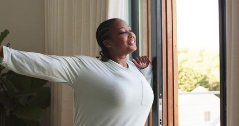 African american woman stretching on sunlit balcony by sliding door wearing casual top