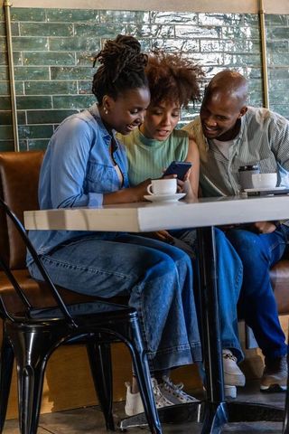 African american friends sharing smartphone and coffee, laughing and smiling in cozy cafe