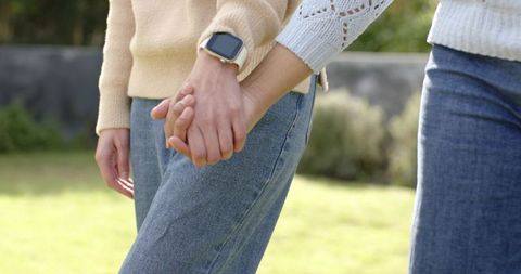 Female partners holding hands wearing knit sweaters and smartwatch walking in sunny park