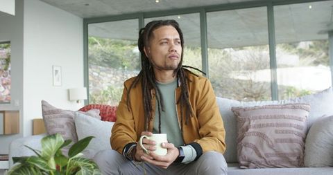 Thoughtful man with dreadlocks holding mug on modern sofa enjoying cozy morning indoors