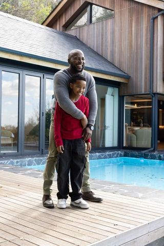 African american father hugging son on wooden deck by modern house and pool