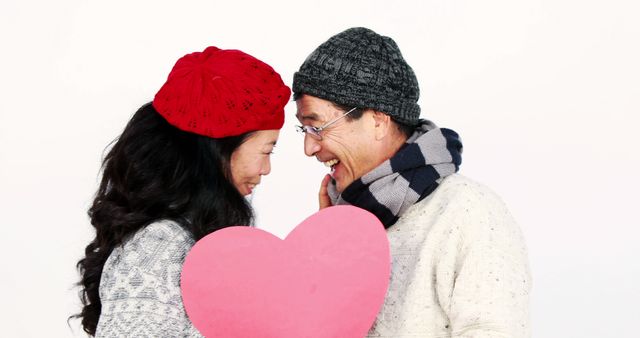 A middle-aged Asian couple shares a joyful moment holding a pink heart together, with copy space. Their warm smiles and winter attire suggest a cozy, affectionate relationship, celebrating a romantic occasion.