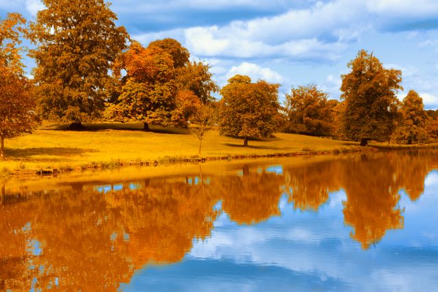 Colorful autumn trees reflected in a serene lake with vibrant foliage and clear blue sky with scattered clouds. Ideal for use in nature-themed websites, travel blogs, seasonal greeting cards, and decorations or wallpaper designs emphasizing tranquility and beauty.