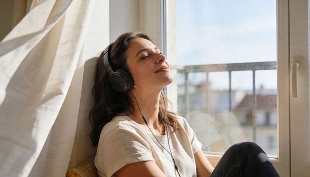 Sunlit woman relaxing with wired headphones by urban apartment window, eyes closed