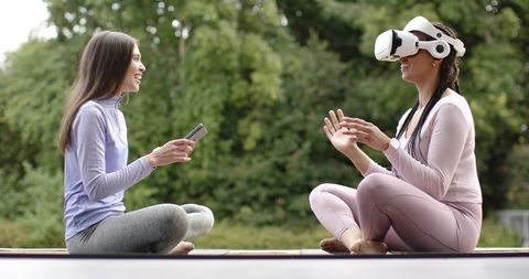 Diverse female friends using vr headset and smartphone while doing outdoor yoga and meditation