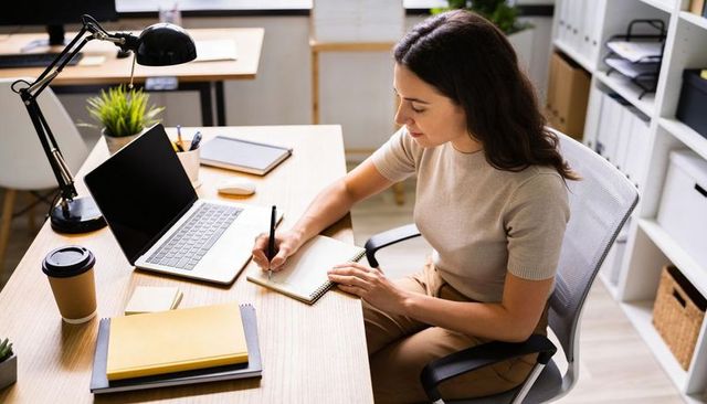 Focused woman writing at modern minimalist desk with laptop and notebook for productivity
