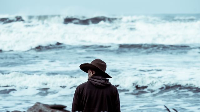 Man wearing a hat is standing by the ocean and looking at rough waves during overcast weather. This image can be used in contexts highlighting themes like solitude, contemplation, adventure, travel, nature, or mental well-being. Useful for blog posts, inspirational quotes, background images, or advertisement purposes.