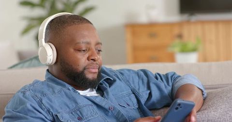 Relaxing african man wearing wireless headphones and scrolling smartphone on couch at home