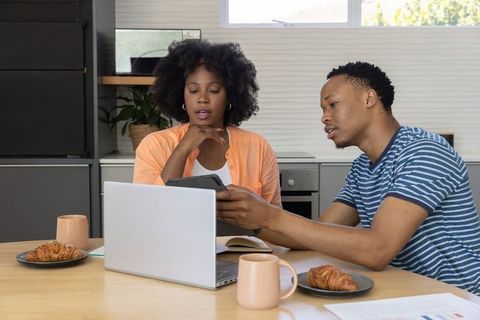 African american couple working from home at kitchen table reviewing tablet and laptop