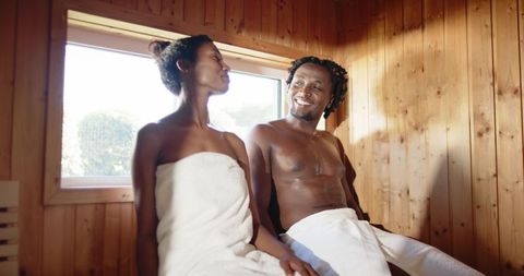 African american couple relaxing in wooden sauna bench with towels and natural daylight