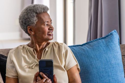 Senior african woman holding smartphone and smiling on sofa in cozy living room