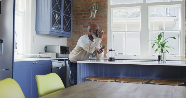 Man enjoying coffee while using a smartphone in contemporary kitchen with ample natural light. Perfect for ads featuring modern lifestyle, kitchen design inspiration, home appliances, or morning routines.