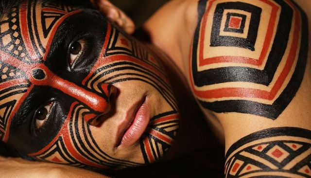 Lying male-presenting model showing geometric tribal face and shoulder body paint closeup