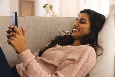 South asian woman relaxing on sofa using smartphone at home