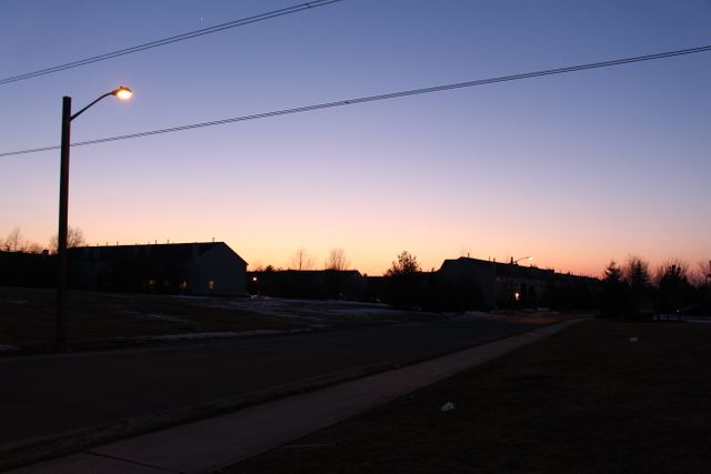 Image depicts a quiet suburban street at dusk, with a lit street light casting a gentle glow. The sky transitions from a gradient of warm colors near the horizon to a darker tone above. Perfect for conveying themes of calm, tranquility, and suburban life. Ideal for use in articles, blogs, and advertisements related to residential areas, urban planning, and peaceful living environments.