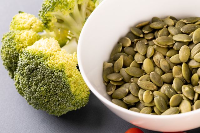 This image showcases a close-up of fresh pumpkin seeds in a white bowl next to a head of broccoli. Ideal for use in articles or blogs about healthy eating, organic food, and nutrition. Perfect for promoting vegan and vegetarian diets, superfoods, and clean eating habits.