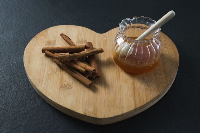 Cinnamon sticks and honey jar with dipper on heart shaped wooden board. Ideal for use in culinary blogs, healthy eating articles, food preparation tutorials, or rustic kitchen decor themes.