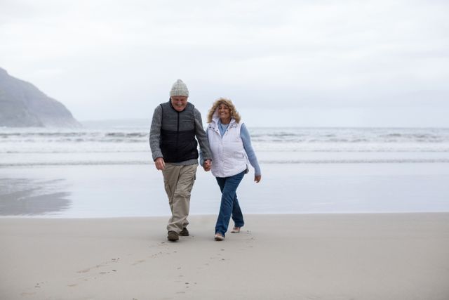 Senior couple enjoying a peaceful walk on the beach, holding hands and smiling. Ideal for use in retirement planning, senior lifestyle, health and wellness, and travel advertisements. Perfect for illustrating themes of love, companionship, and active aging.