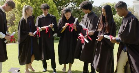 Diverse graduates celebrating with diplomas tied with red ribbons on sunny campus lawn