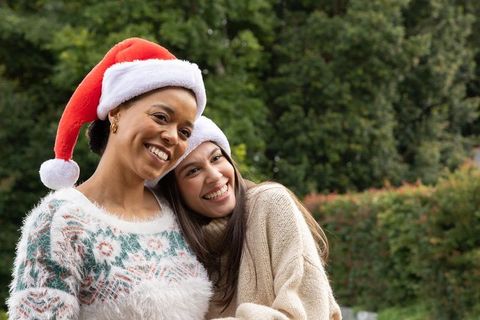 Two diverse women wearing santa hats smiling in cozy knit sweaters outdoors for holiday