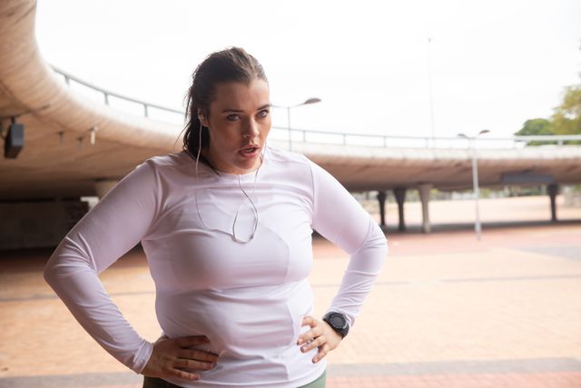 Curvy Caucasian woman with long dark hair wearing sports clothes and earphones exercising in a city, standing with hands on hips, taking a rest and cooling off during her workout