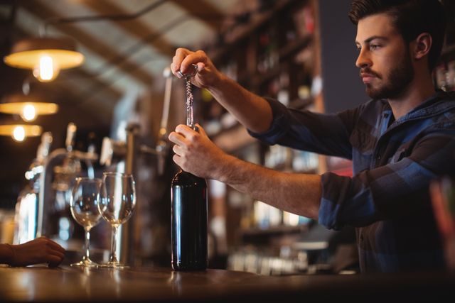 Male bartender using corkscrew to open wine bottle at bar counter. Wine glasses ready for serving. Ideal for illustrating bar service, hospitality industry, nightlife scenes, or wine-related content.