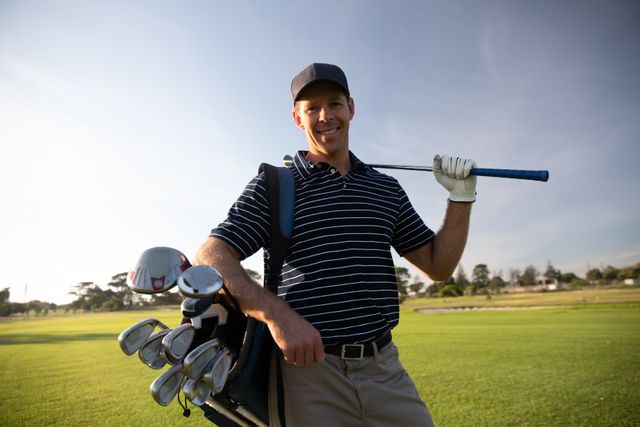 Smiling male golfer posing on a sunny day at a golf course, holding a golf club on his shoulder and carrying a golf bag. Ideal for advertisements, brochures, or articles related to golfing, outdoor activities, healthy lifestyles, and leisure sports. Perfect for promoting golf equipment, apparel, or courses.