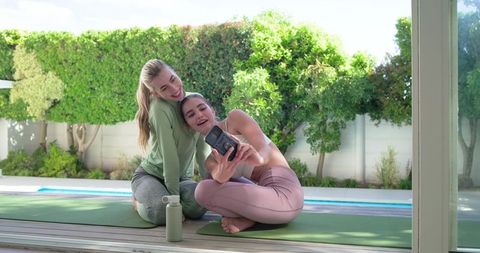 Friends taking instant-camera selfie after morning yoga on sunlit poolside deck, smiling