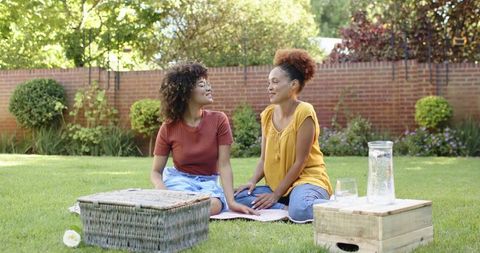 Diverse women chatting on picnic blanket in sunny backyard relaxing with basket, pitcher