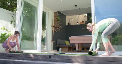 Two women rolling out yoga mats on modern glass patio for outdoor morning yoga practice