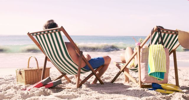 This image shows a couple relaxing on striped deck chairs on a sandy beach, enjoying an ocean view. Ideal for travel blogs, vacation advertisements, relaxation and leisure themes. Perfect for summer holiday promotions, lifestyle magazines, and tourism websites.