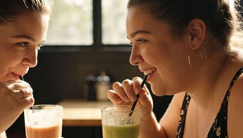 Two women laughing and sipping green and pink smoothies in cozy sunlit coffeehouse