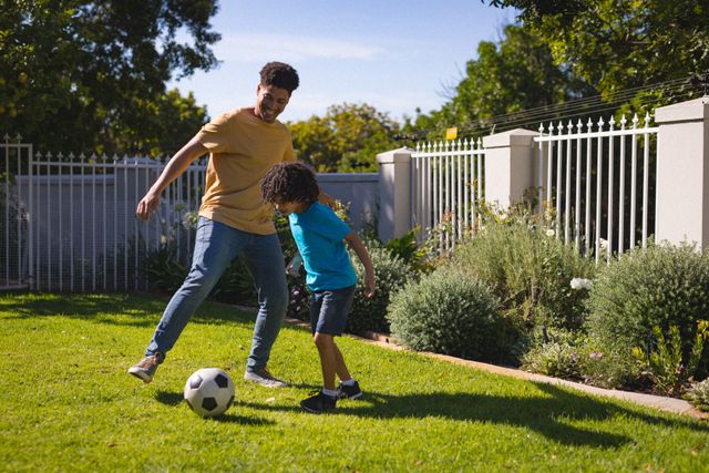This image depicts a Hispanic father and son enjoying a game of soccer in their backyard on a sunny day. The father is engaging with his son, creating a moment of bonding and fun. This photo can be used for promoting family activities, parenting blogs, lifestyle articles, and advertisements focusing on outdoor sports and family togetherness.