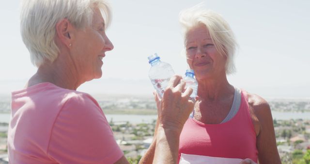 Senior diverse women wearing pink t-shirts holding bottles and drinking water. Retirement, friendship, healthy and active lifestyle.