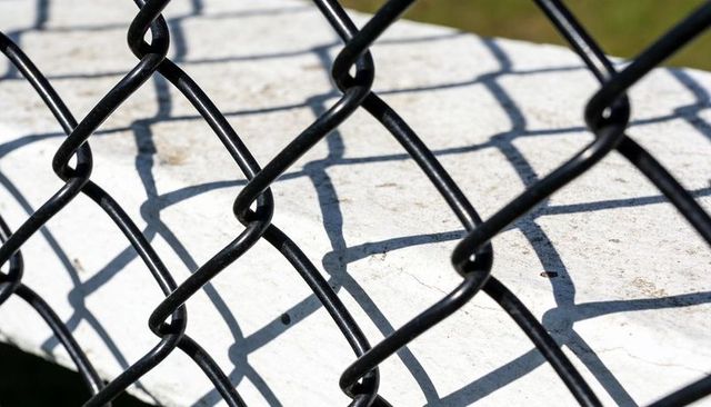Black chain-link fence casting diamond shadows on sunlit concrete ledge, abstract texture