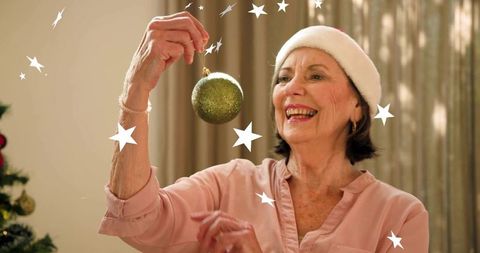 Senior woman holding green christmas ornament smiling in cozy holiday home wearing santa hat