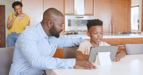 African American Father Guiding Son Using Tablet at Kitchen While Mother Talking on Phone