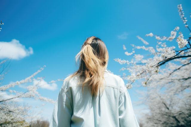 Woman standing amidst blossoming trees under a clear, sunny sky. Ideal for spring promotion materials, nature blogs, relaxation themes, wellness content, and outdoor activity features.