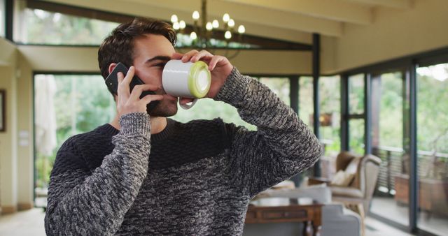 Scene depicts a man engaging with both a coffee cup and smartphone, illustrating multitasking and modern living. Bright, cozy room contributes to a sense of warmth and comfort. Useful for lifestyle blogs, articles about work-life balance, advertisements showing relaxed home environments, and technology or communication themes.