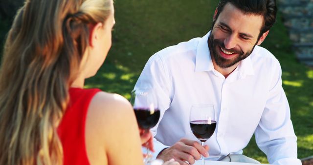A Caucasian man and woman enjoy a conversation over glasses of red wine in an outdoor setting, with copy space. Their casual yet intimate interaction suggests a romantic or friendly gathering on a pleasant day.