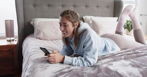 Mid-adult woman lounging on bed with smartphone in cozy sunlit bedroom wearing hoodie