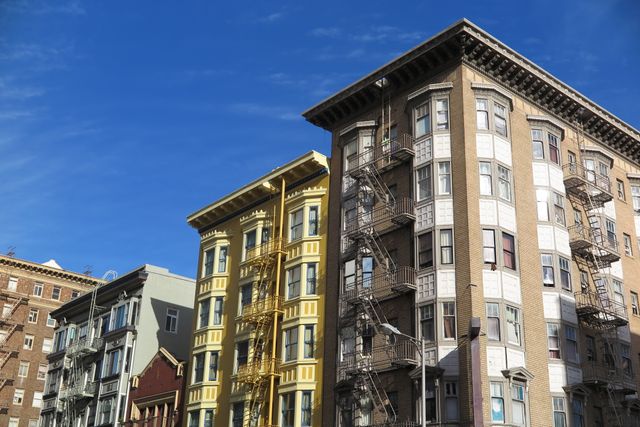 This image captures a row of historic urban buildings with fire escapes under a bright blue sky. Ideal for use in presentations, articles, or websites about city living, architecture, and urban development. Perfect for depicting downtown environments or residential areas with classic charm.