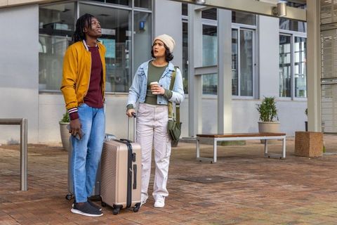 Young diverse travelers waiting with rolling luggage and coffee in urban plaza