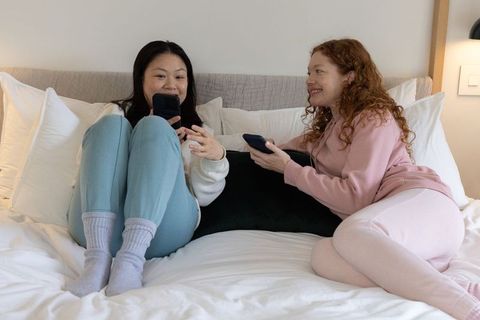 Two women relaxing on bed in loungewear, scrolling phones and chatting in cozy bedroom