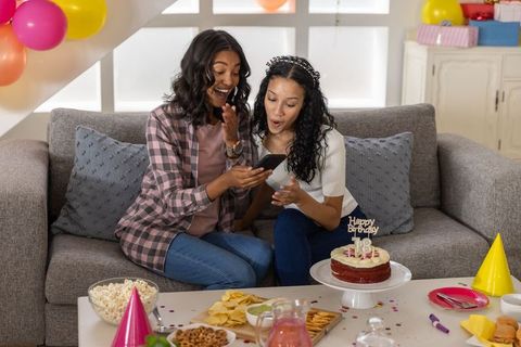Diverse sisters celebrating 18th birthday on cozy gray sofa with cake and balloons