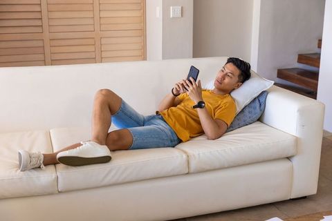 Asian man lounging on modern white sofa scrolling smartphone wearing smartwatch in casual outfit