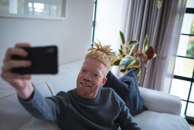 Albino African American man sitting on a white couch in a modern living room, taking a selfie with his smartphone. He is smiling and appears relaxed, enjoying leisure time at home. This image can be used for articles or advertisements related to technology, modern lifestyle, home relaxation, or diversity and inclusion.