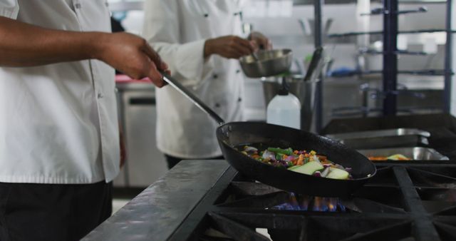 Two chefs wearing white uniforms are cooking vegetables on a stove in a professional kitchen. The image shows one chef holding a frying pan with sautéed vegetables while another chef operates in the background. Ideal for illustrating culinary training, restaurant advertisements, or articles about professional cooking and culinary arts.