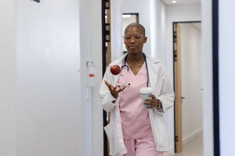 African clinician walking clinic corridor tossing apple while holding coffee and stethoscope